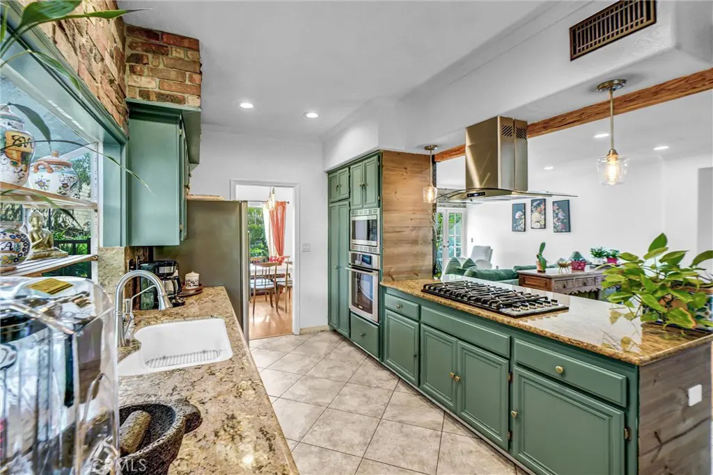 A bright kitchen with green cabinets, granite countertops, and stainless steel appliances. A dining area and living room are visible in the background.