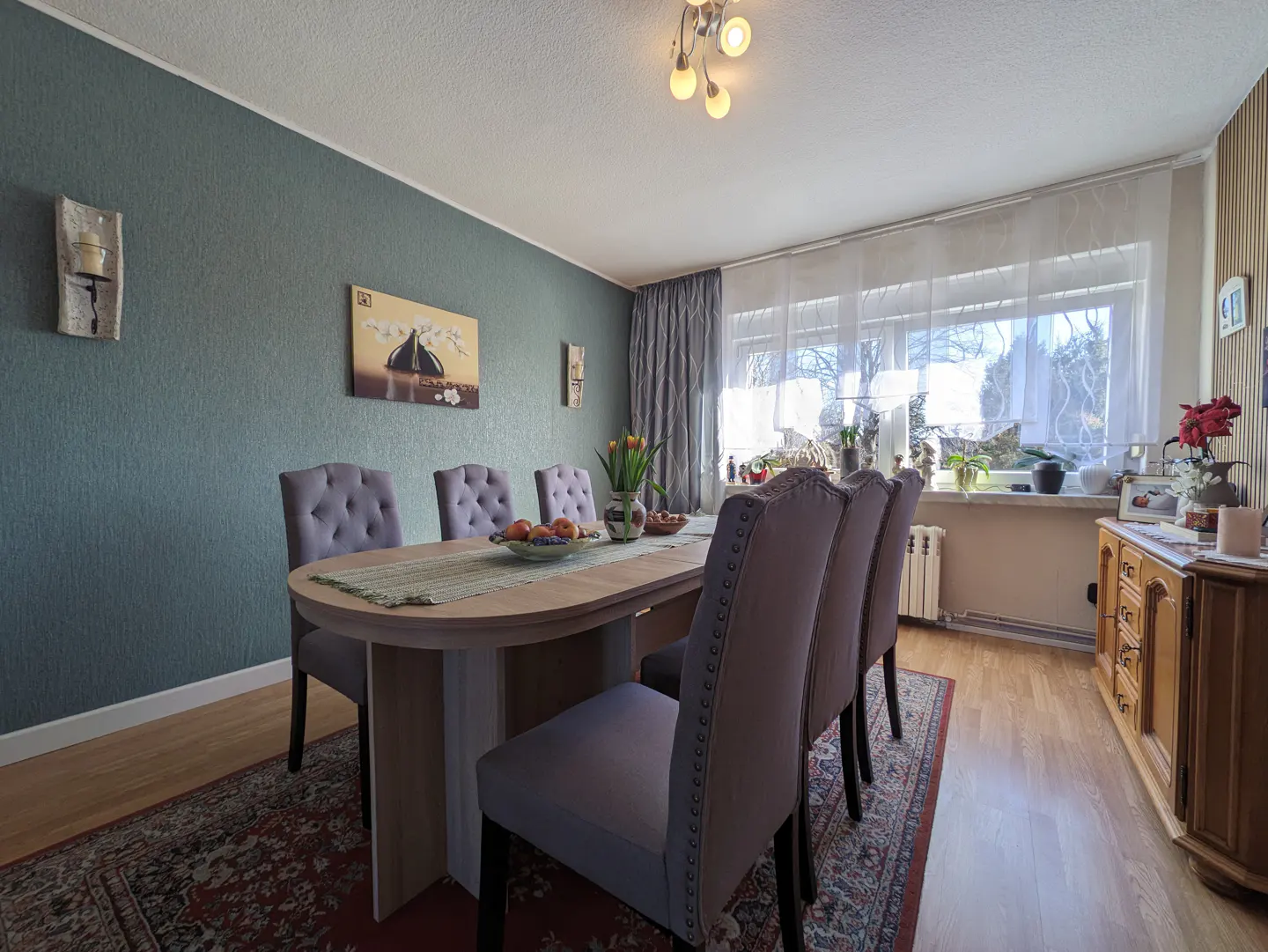 Dining room with oval table, six gray chairs, and a red patterned rug. Teal walls, wood floors, and a window with sheer curtains.