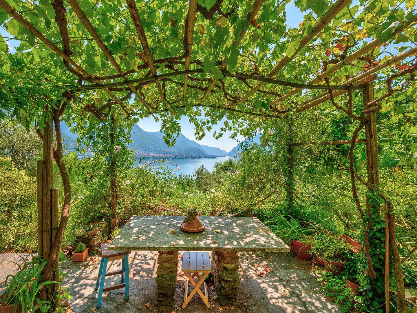 Outdoor dining area with stone table and chairs under a vine-covered pergola, overlooking a lake and mountains. Lush greenery surrounds the space.