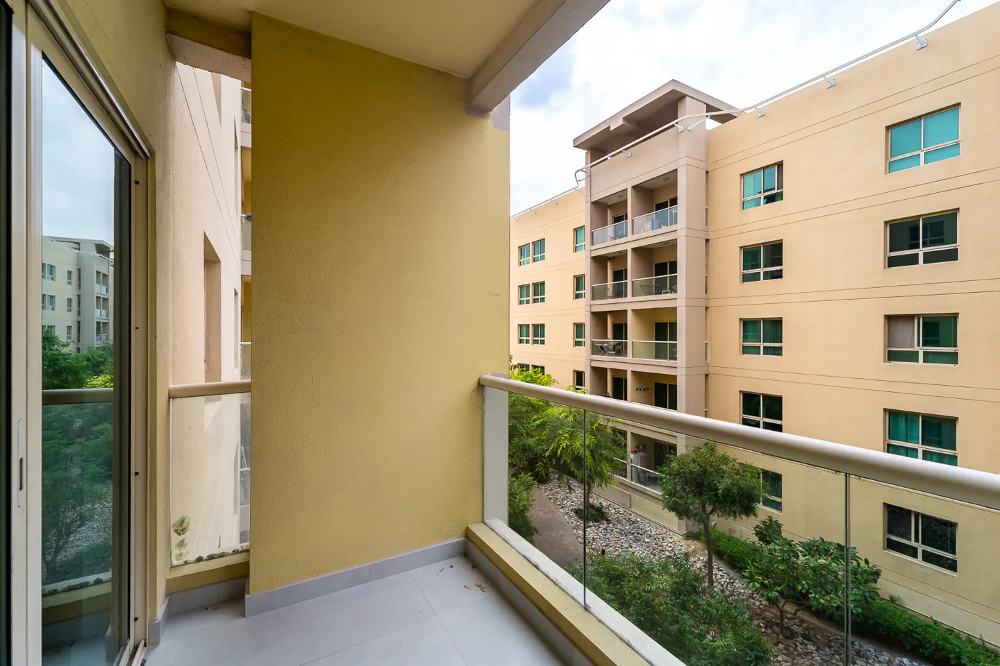 View from a balcony with glass railings, overlooking a courtyard with trees and a beige apartment building.