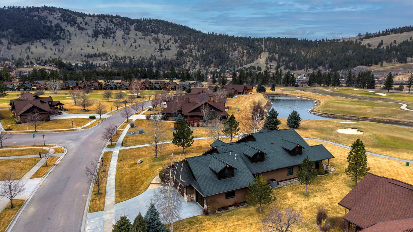 Aerial view of a brown house with a green roof, a golf course, a pond, and mountains in the background.