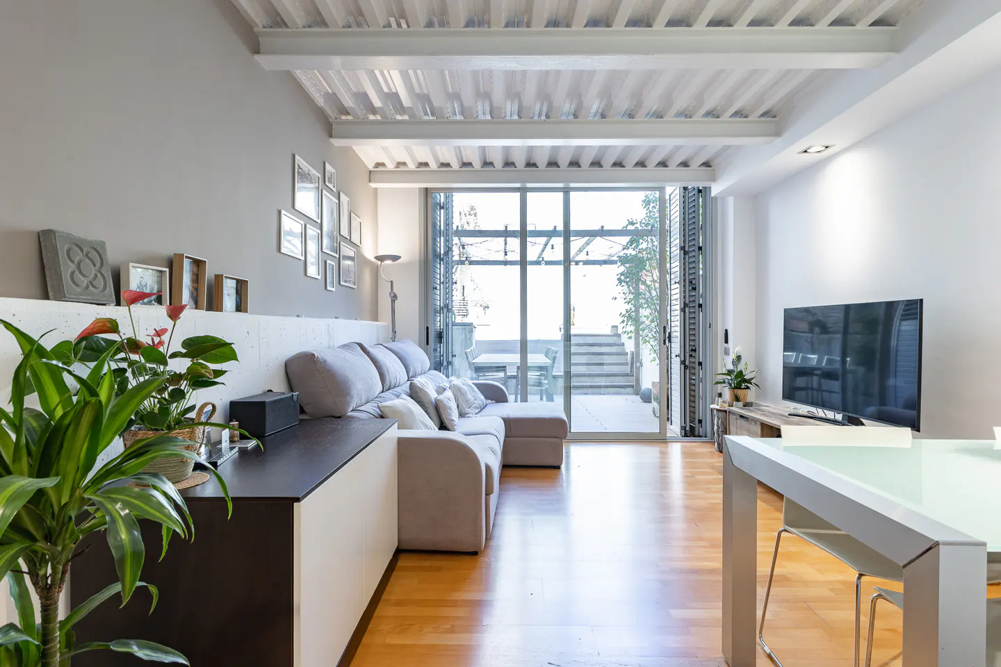 Living room with gray sofa, wood floors, and sliding glass doors to a patio. White ceiling with exposed beams. TV and dining table visible.