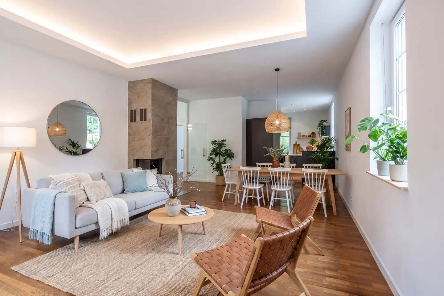 Bright, open-concept living and dining area with a gray sofa, round mirror, and a dining table with white chairs. Two brown leather chairs in the foreground. Hardwood floors and white walls.
