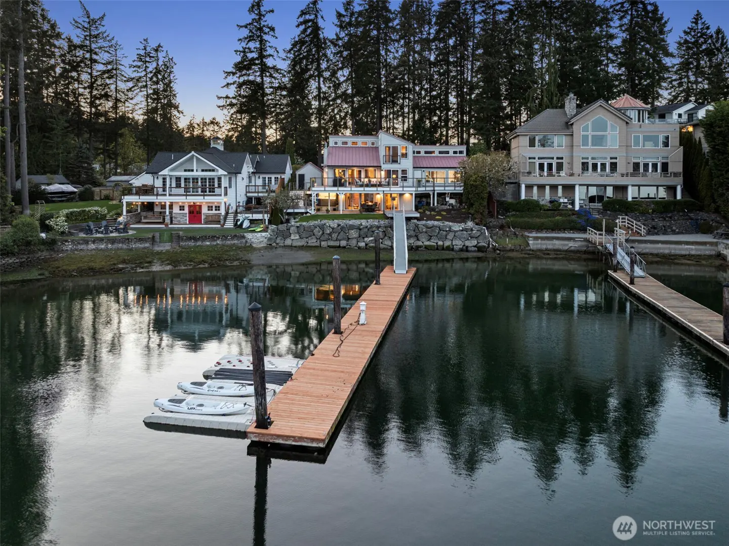 Waterfront homes with docks and kayaks on calm water, reflecting lights. Trees in the background.