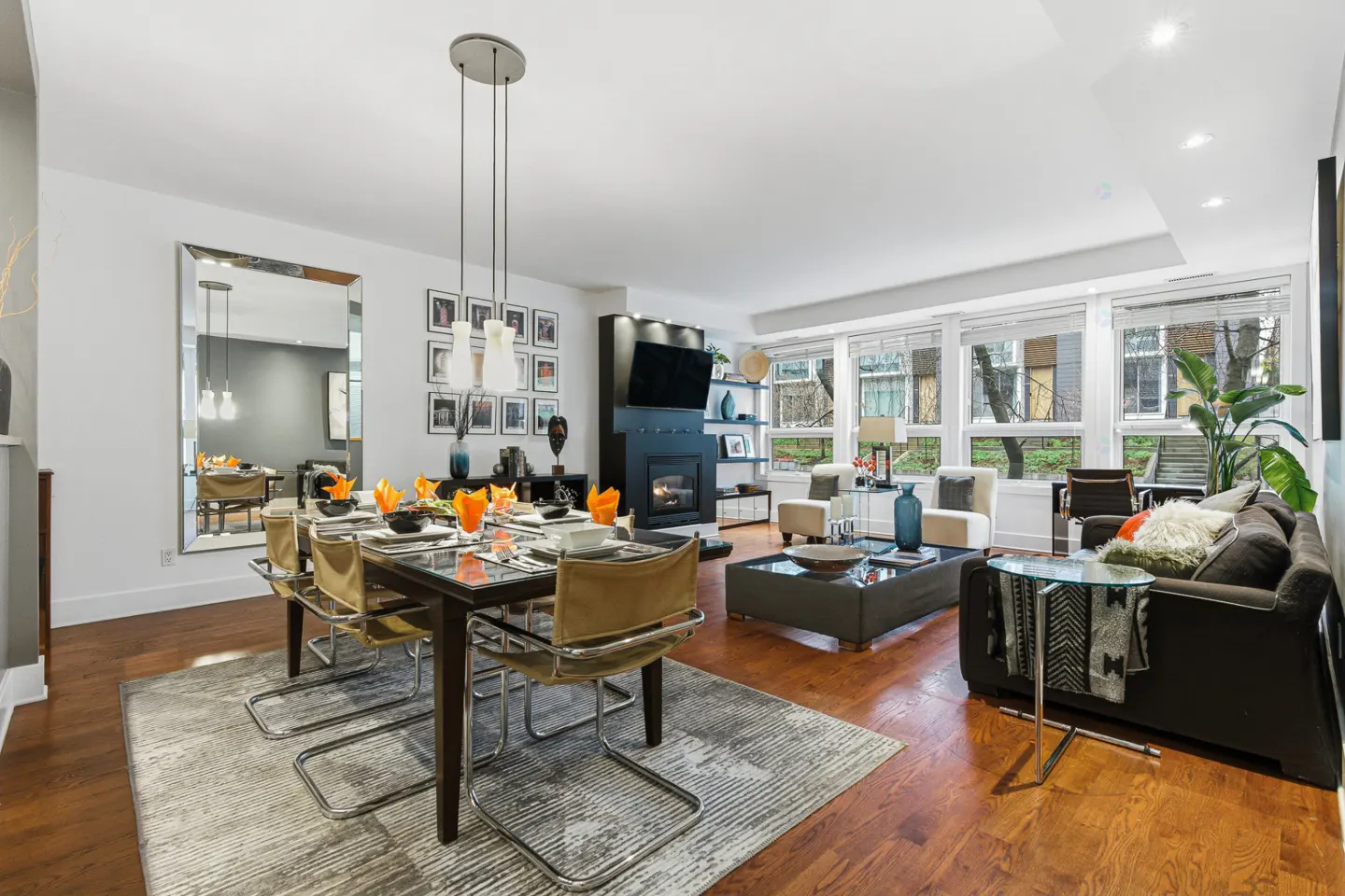 Open-concept living and dining area with hardwood floors, a fireplace, and large windows. Dining table set for a meal.