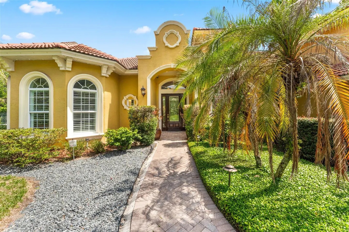 Front view of a yellow stucco house with a red tile roof, palm trees, and a stone walkway.