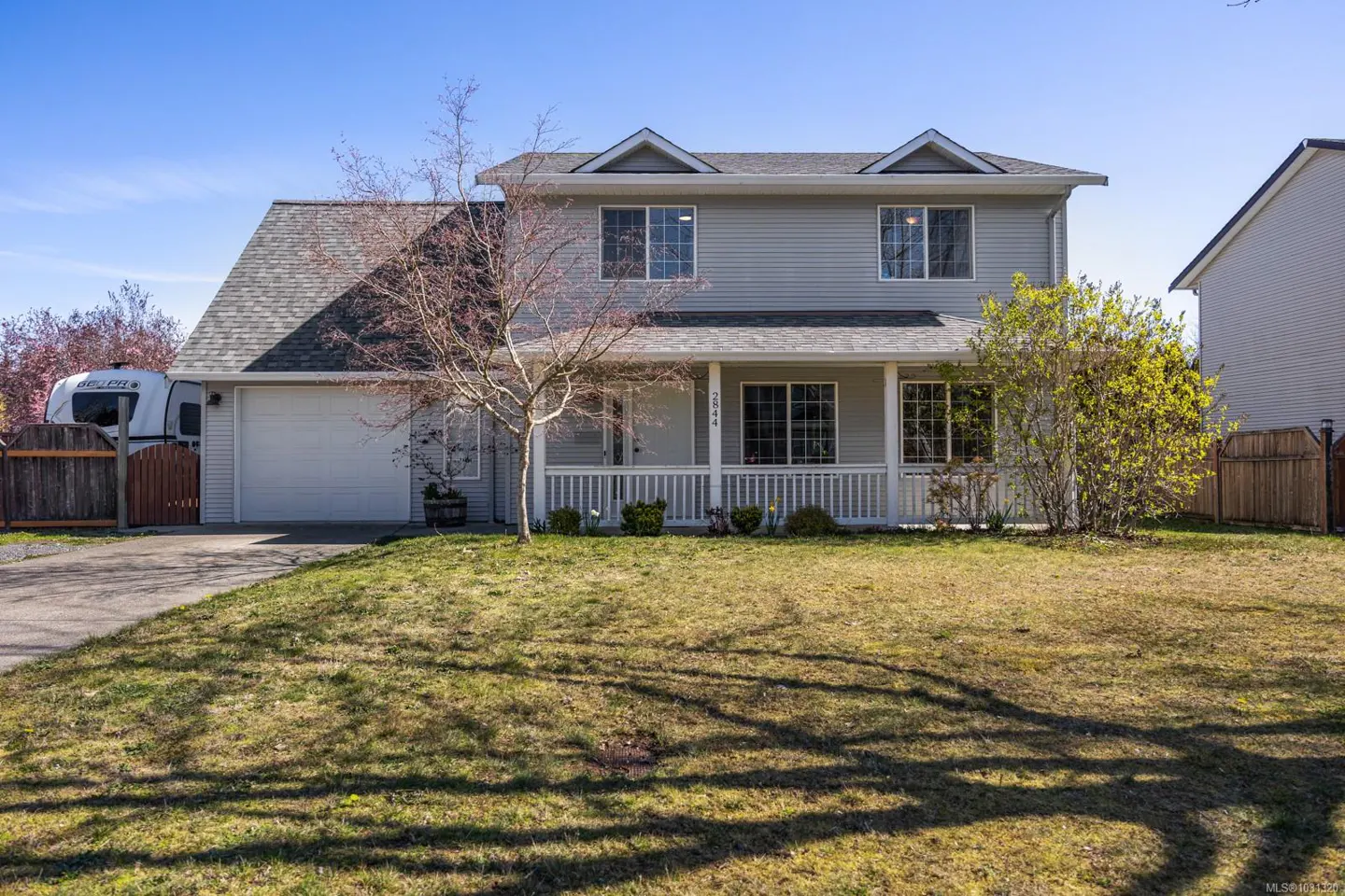 Two-story house with gray siding, a front porch, and a gray roof on a sunny day. A camper is parked in the driveway.