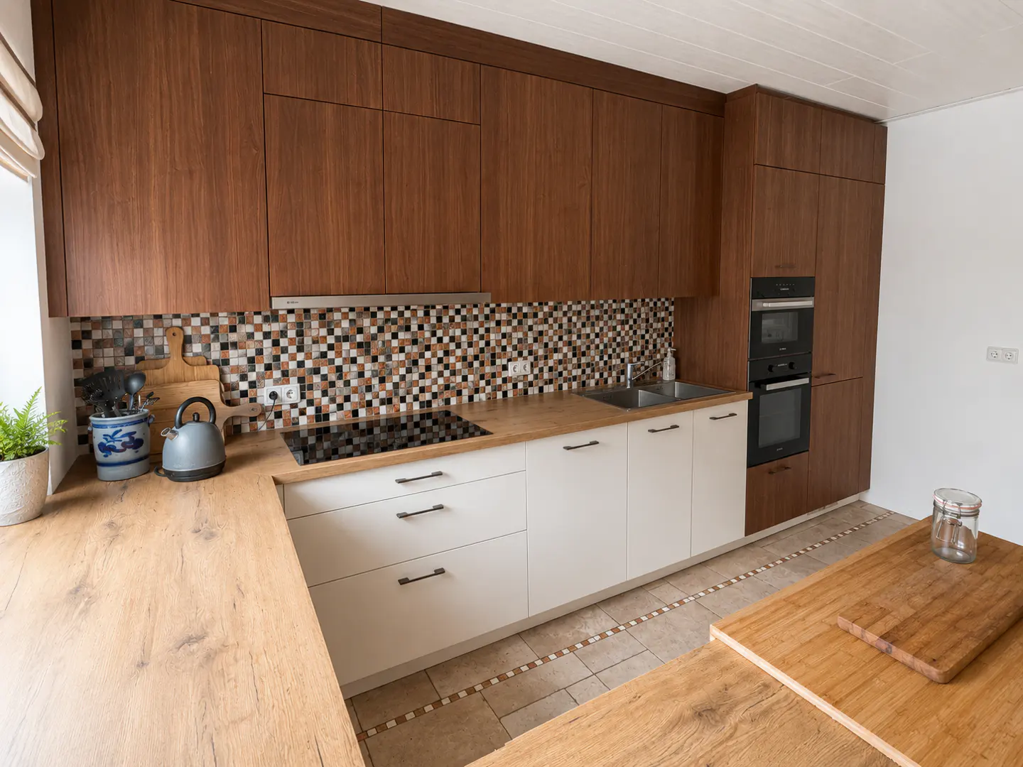A modern kitchen with brown cabinets, white lower cabinets, and a mosaic tile backsplash. A wooden countertop and island add warmth.