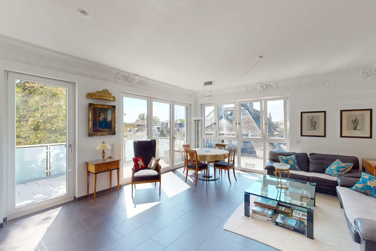 Bright living room with gray tile floor, white walls, and large windows. Features a dining table, sofa, and decorative art.