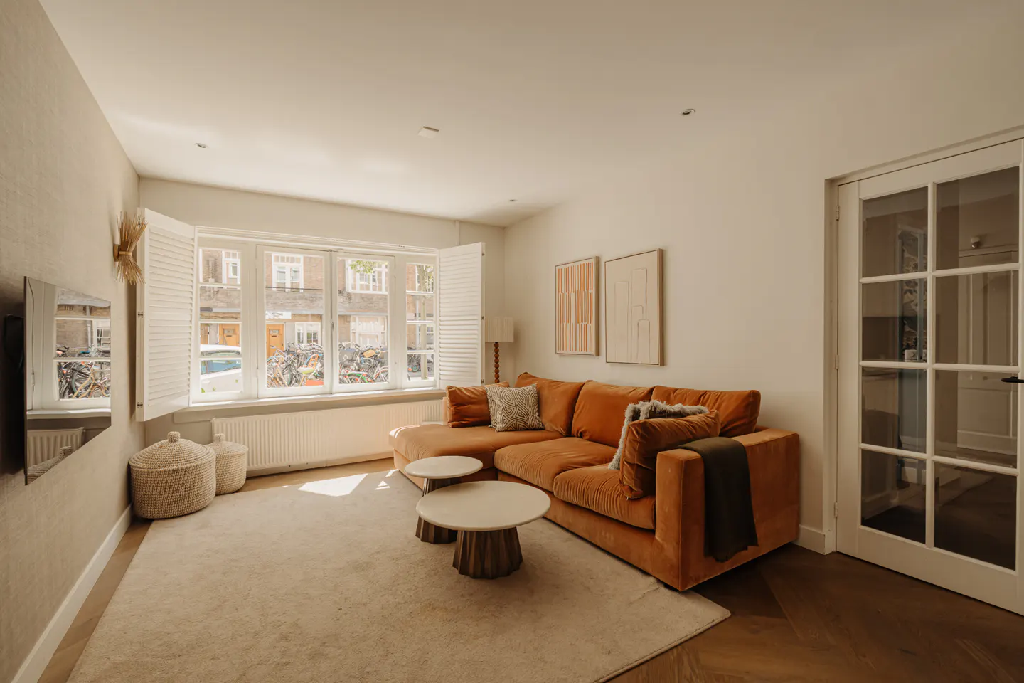 Warm living room with orange sectional sofa, two round tables, and a large window with white shutters. Neutral rug and hardwood floors.