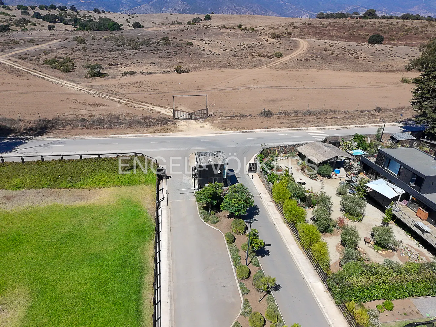 Aerial view of a gated community entrance with green lawns, trees, and houses, set against a dry, hilly landscape.