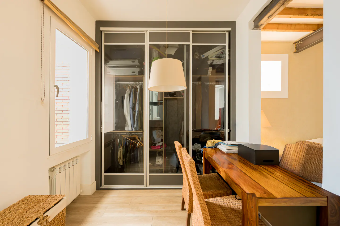 Interior view of a room with a wooden table, chairs, and a closet with sliding glass doors. A white lamp hangs above the table.