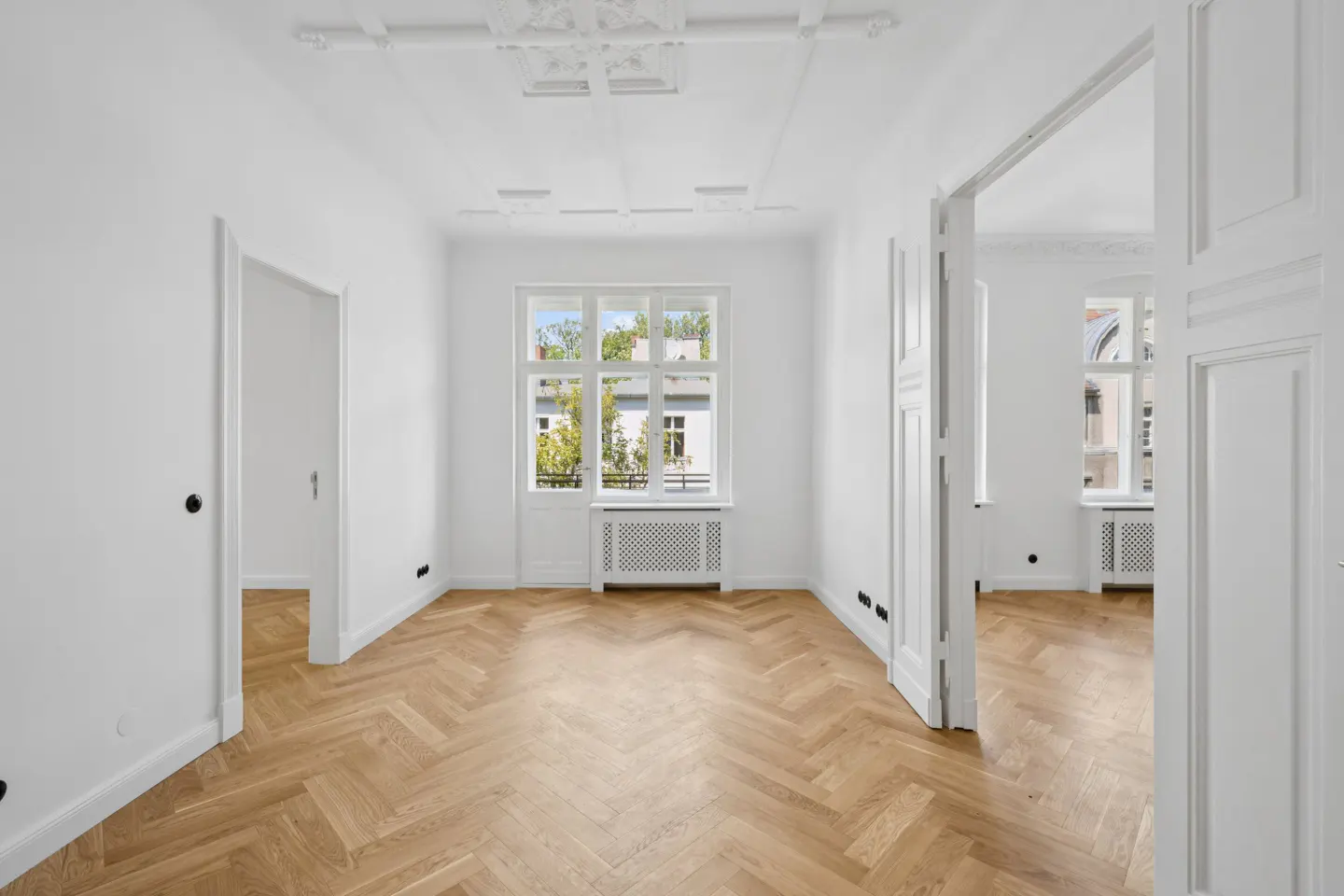 Bright, empty room with white walls, ornate ceiling molding, herringbone wood floors, and large windows overlooking trees.