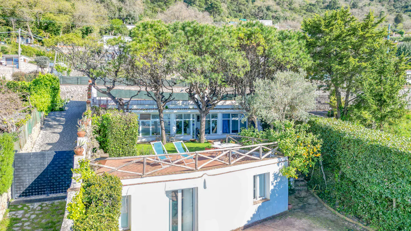 Aerial view of a white villa with a rooftop terrace, two blue lounge chairs, and lush green trees.
