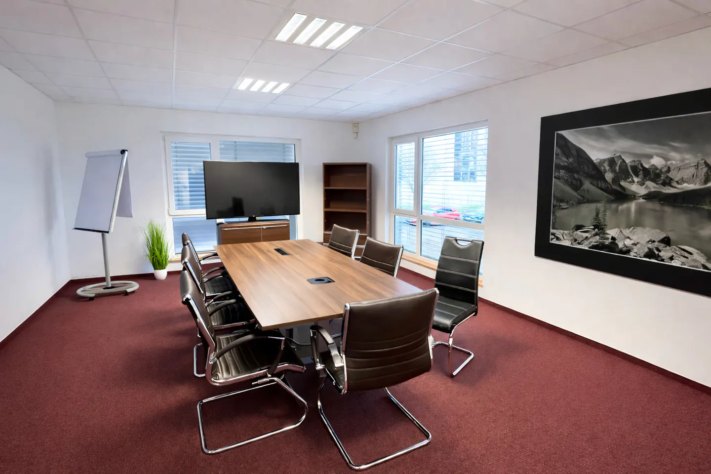 A conference room with a long wooden table, brown leather chairs, and a large TV. A whiteboard and a mountain landscape print are also visible.