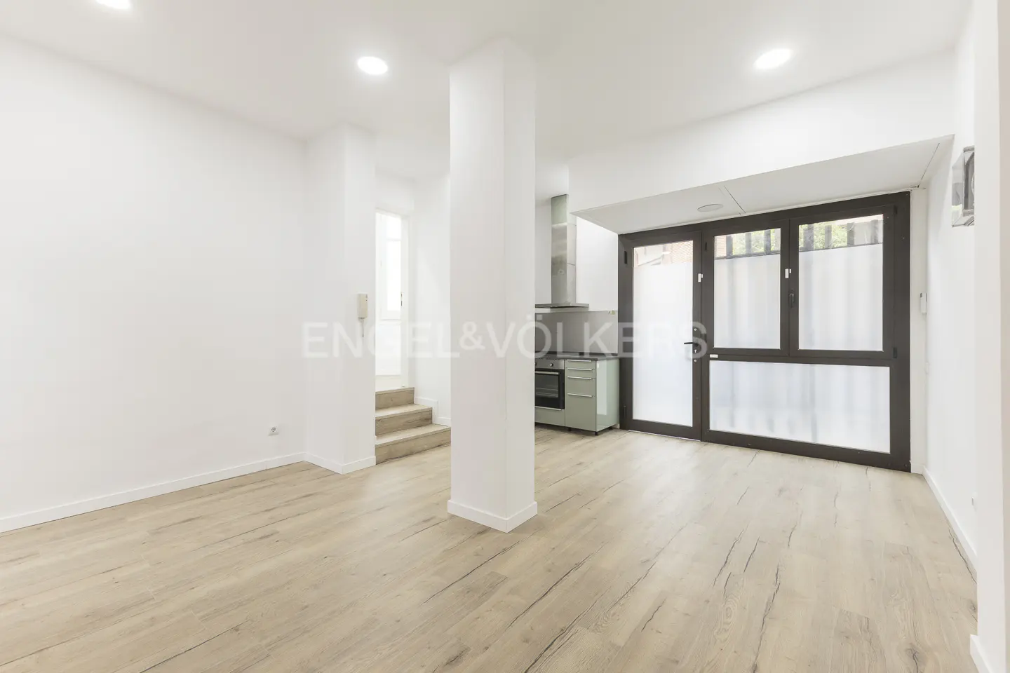 Bright, modern apartment interior with light wood floors, white walls, and a sage green kitchen visible in the background.