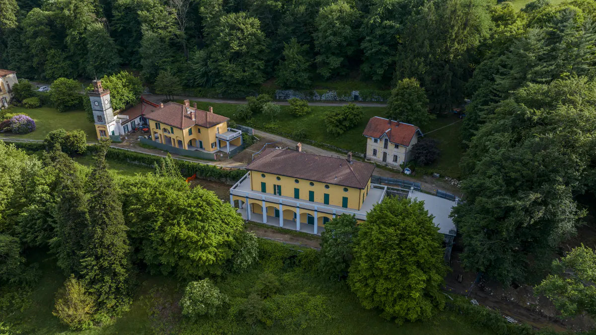 Aerial view of yellow buildings with red roofs, surrounded by green trees and grass. A clock tower is visible on the left.