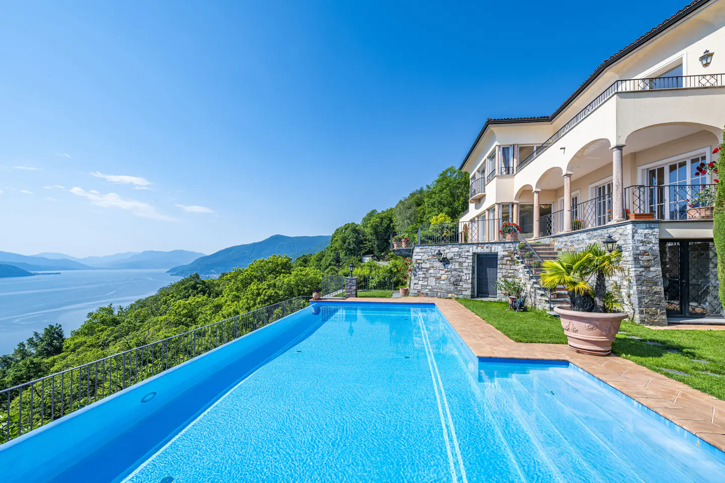 Infinity pool overlooking a lake and mountains next to a luxury villa with stone walls and a sunny terrace.