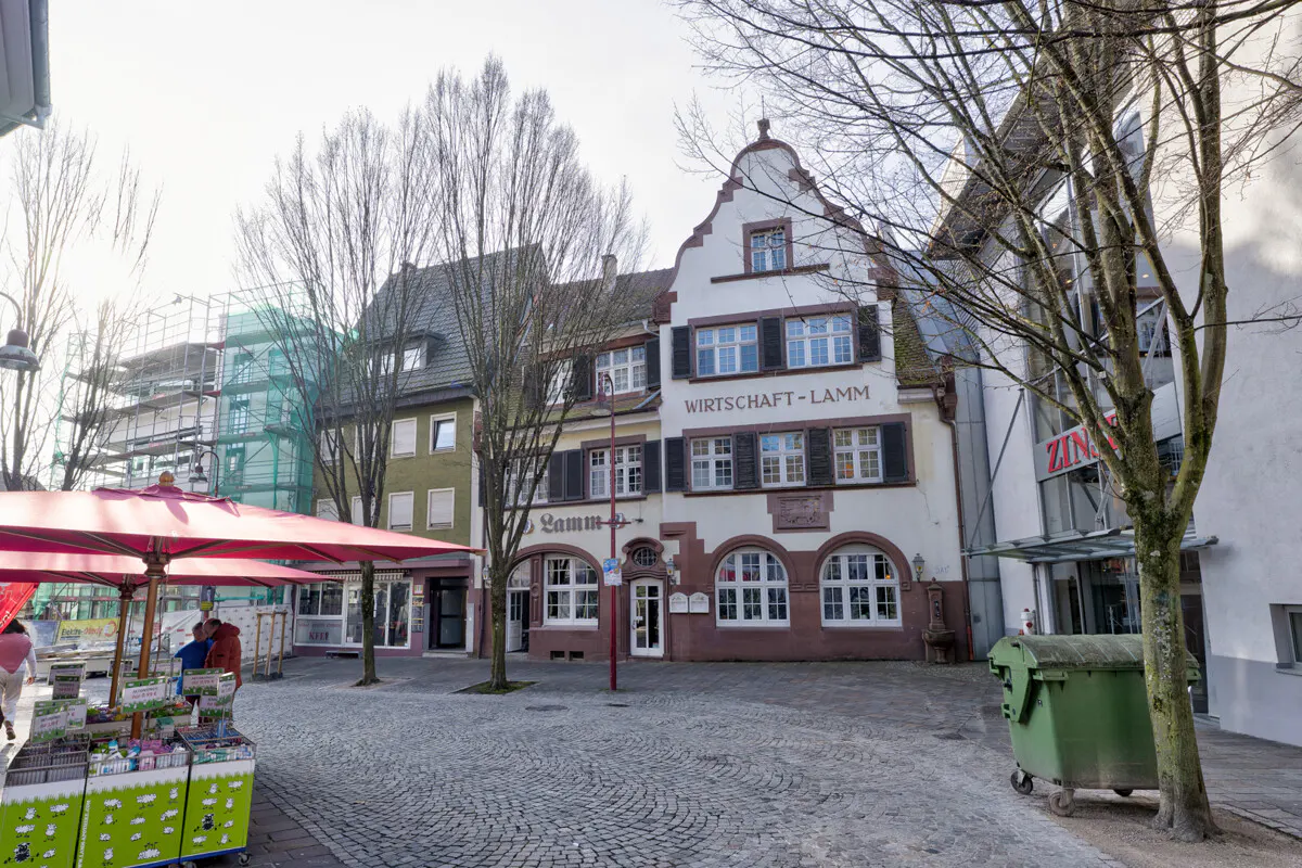 Cobblestone square with a market stall and the "Wirtschaft-Lamm" building in the background. Bare trees and a green dumpster are visible.