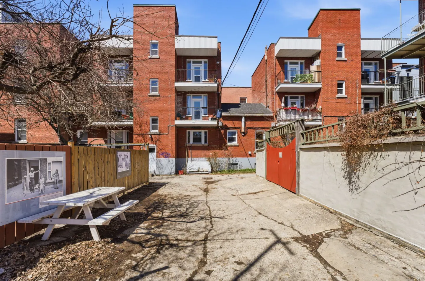 Exterior view of a red brick apartment building with balconies, a picnic table, and a red gate.
