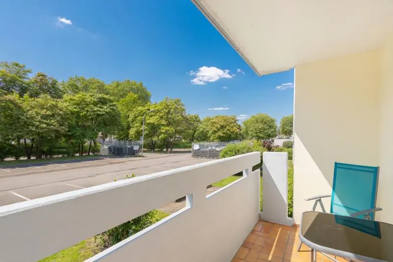 Balcony view with blue chair and table. White railing overlooks parking lot and trees under a blue sky.
