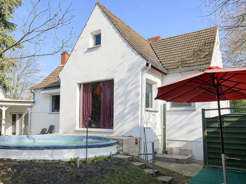Backyard view of a white house with a pool, red umbrella, and green fence. The house has a brown roof and a large window with red curtains.