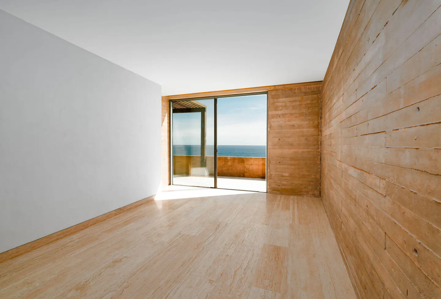 Minimalist room with travertine floors, white wall, and textured brown wall. Sliding glass doors open to a balcony with ocean view.