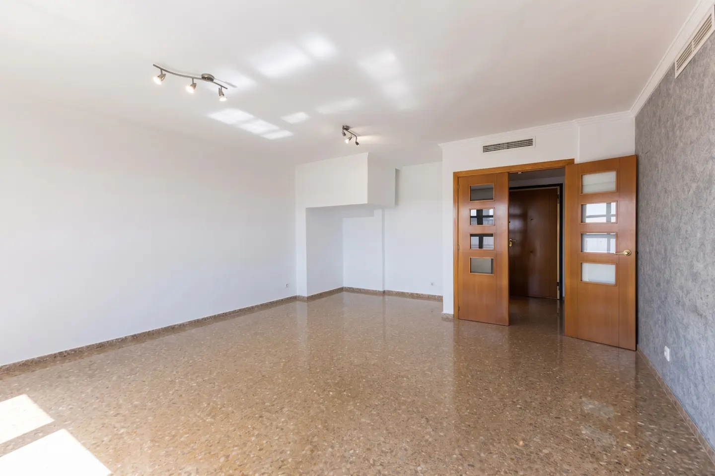 Empty room with white walls, speckled brown tile floors, modern ceiling lights, and open wooden doors to a hallway.