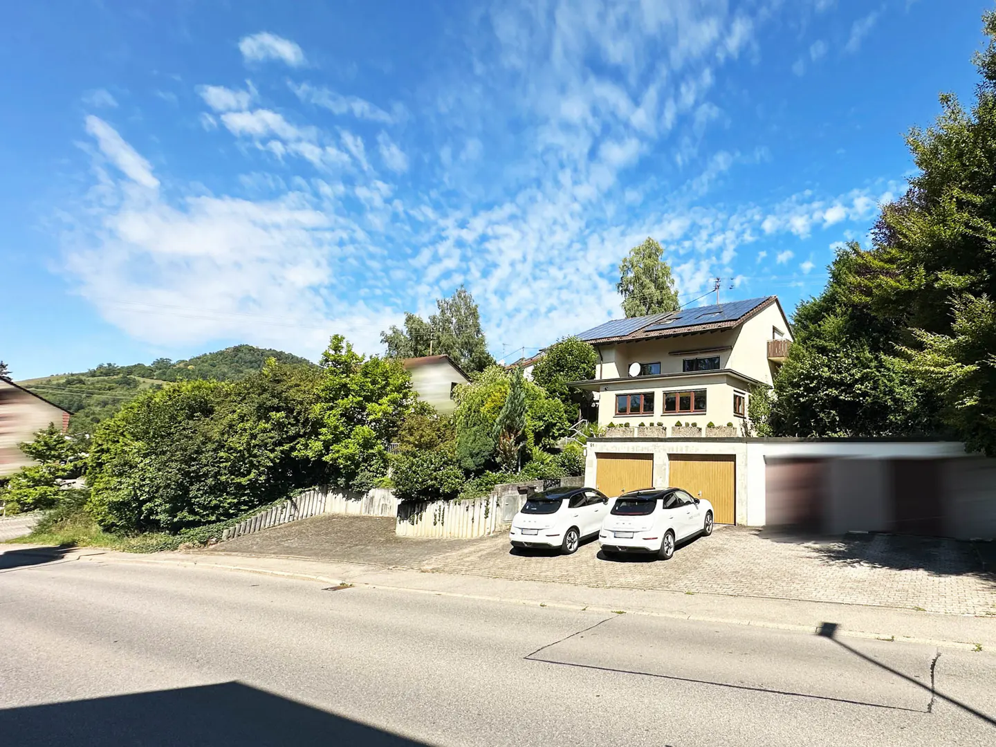 Two-story house with solar panels, two cars in driveway, and lush greenery under a blue, cloudy sky.