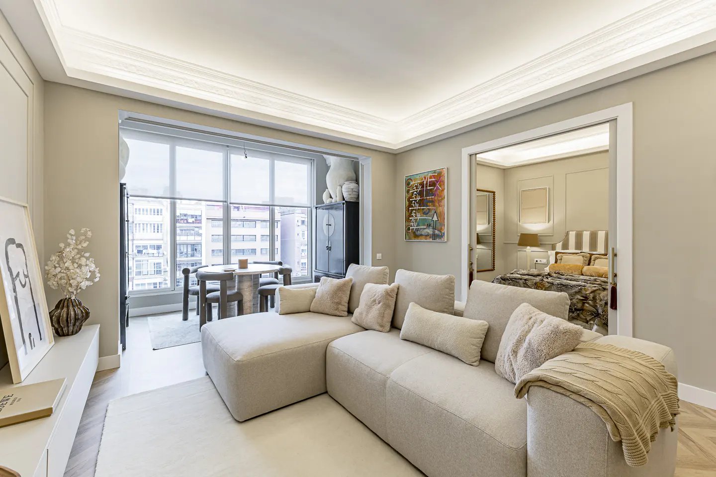 Bright living room with beige sectional sofa, pillows, and throw blanket. Dining table by window with city view. Bedroom visible through doorway.