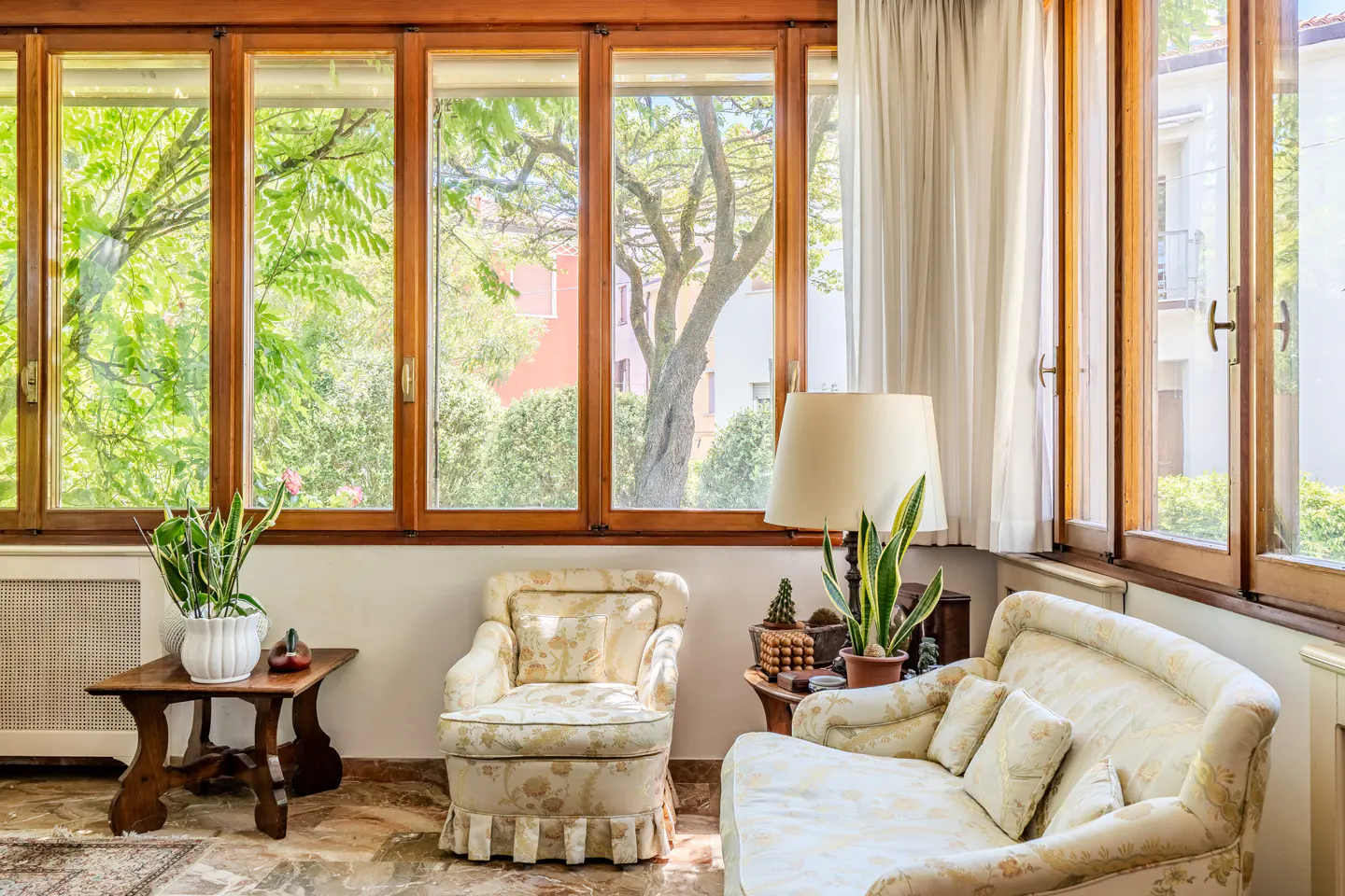 Bright sunroom with wood-framed windows overlooking green trees. Floral armchair and sofa with a lamp and plants.