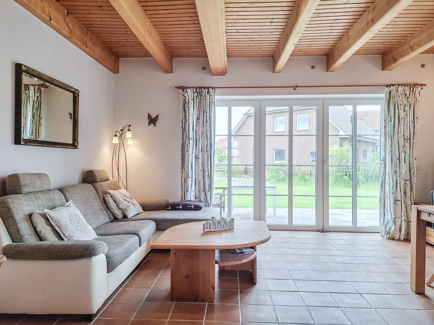Living room with exposed wood beams, tile floor, and large glass doors to the outside. A gray sofa and wood coffee table are in the foreground.