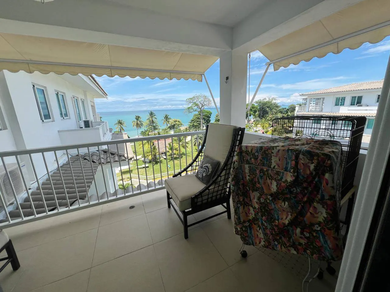Balcony view with chair, table, and ocean. White railings and awnings frame the scene. Palm trees and blue water visible in the background.