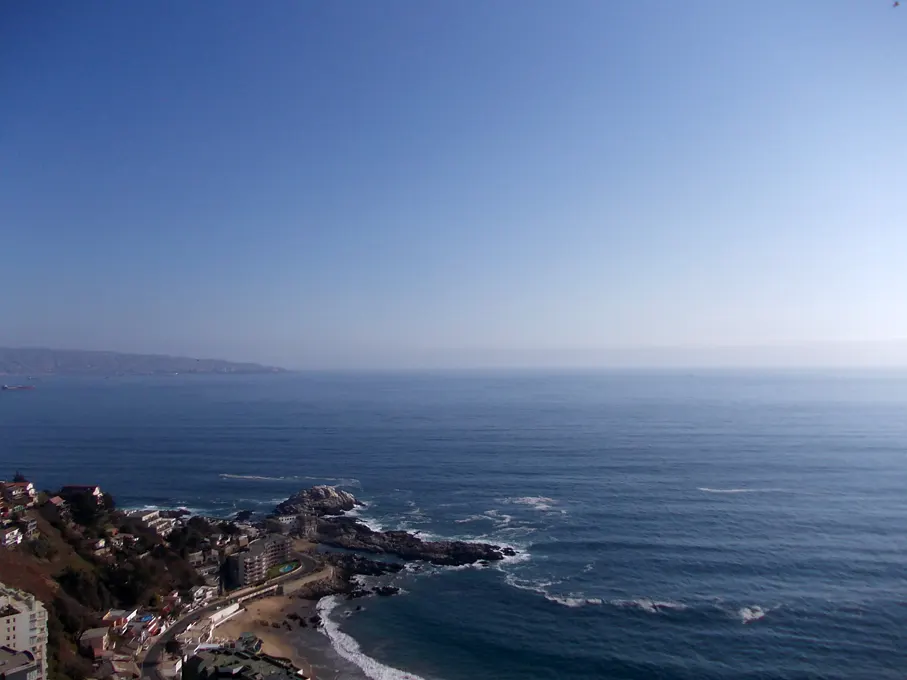 Ocean view with blue sky, waves crashing on rocks, and buildings on the coastline.