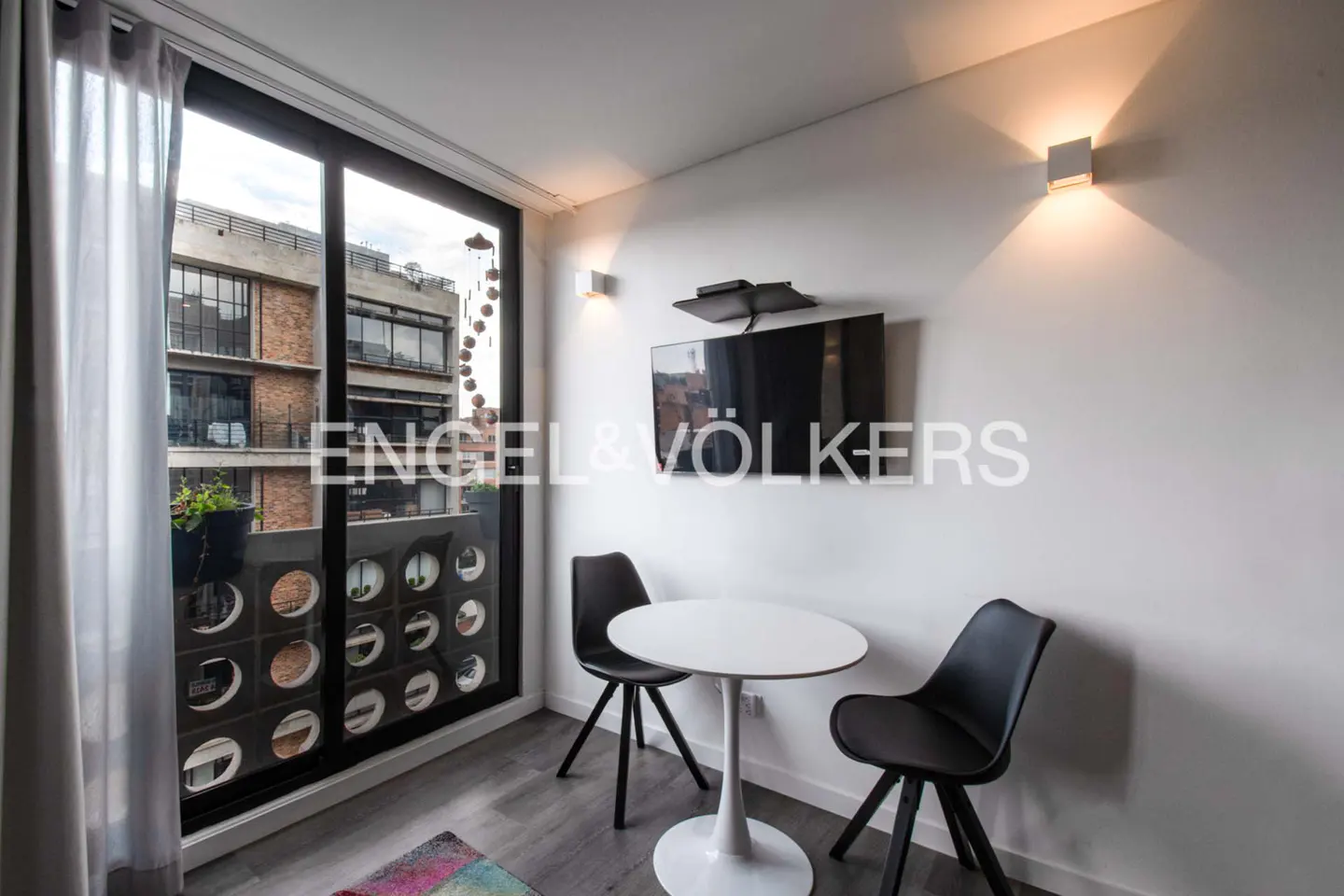 A modern apartment interior with a white table, two black chairs, and a TV on the wall. A large window shows a city view.