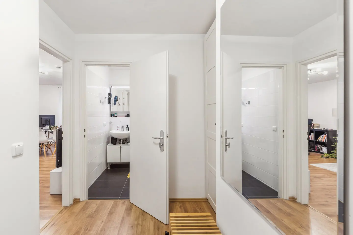 Hallway with wood floors, white walls, and open doors to a bathroom and living area. A mirror reflects the bathroom and living area.