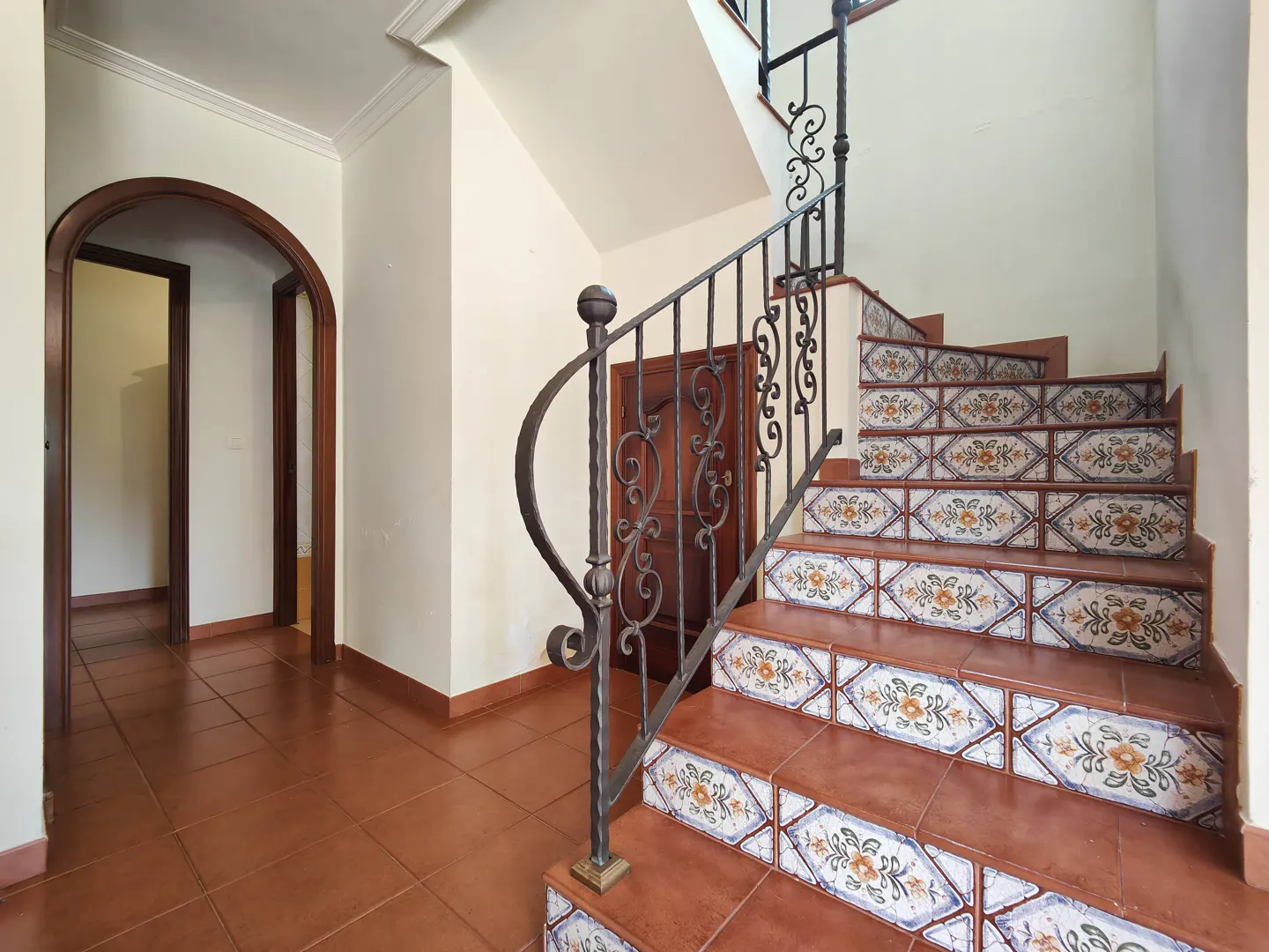 Interior view of a home featuring a staircase with decorative tile risers and a wrought iron railing. Brown tile flooring and arched doorways are visible.