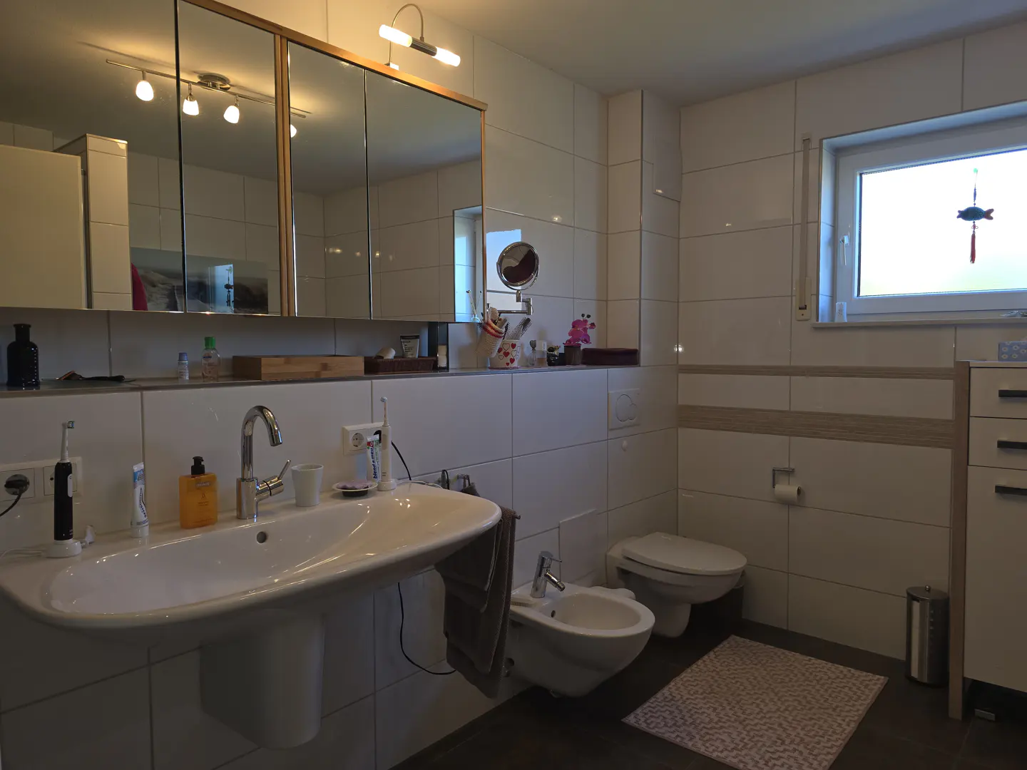 Bright bathroom with white tile walls, a large sink, toilet, and bidet. A window lets in natural light.