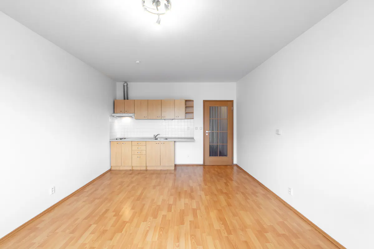 Empty room with light wood flooring, white walls, and a small kitchenette with light wood cabinets and a glass-paneled door.