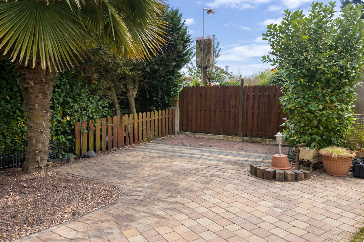A brick-paved patio with a palm tree, wooden fences, and green bushes under a partly cloudy sky.