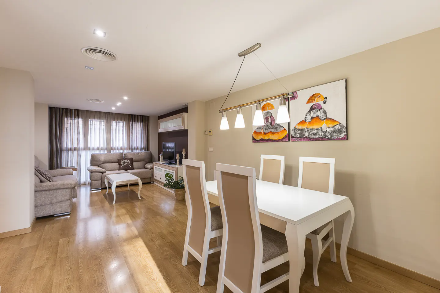 A bright, open-concept living and dining area with wood floors. A white dining table with four chairs sits near a wall with artwork. A gray sofa and TV are in the background.