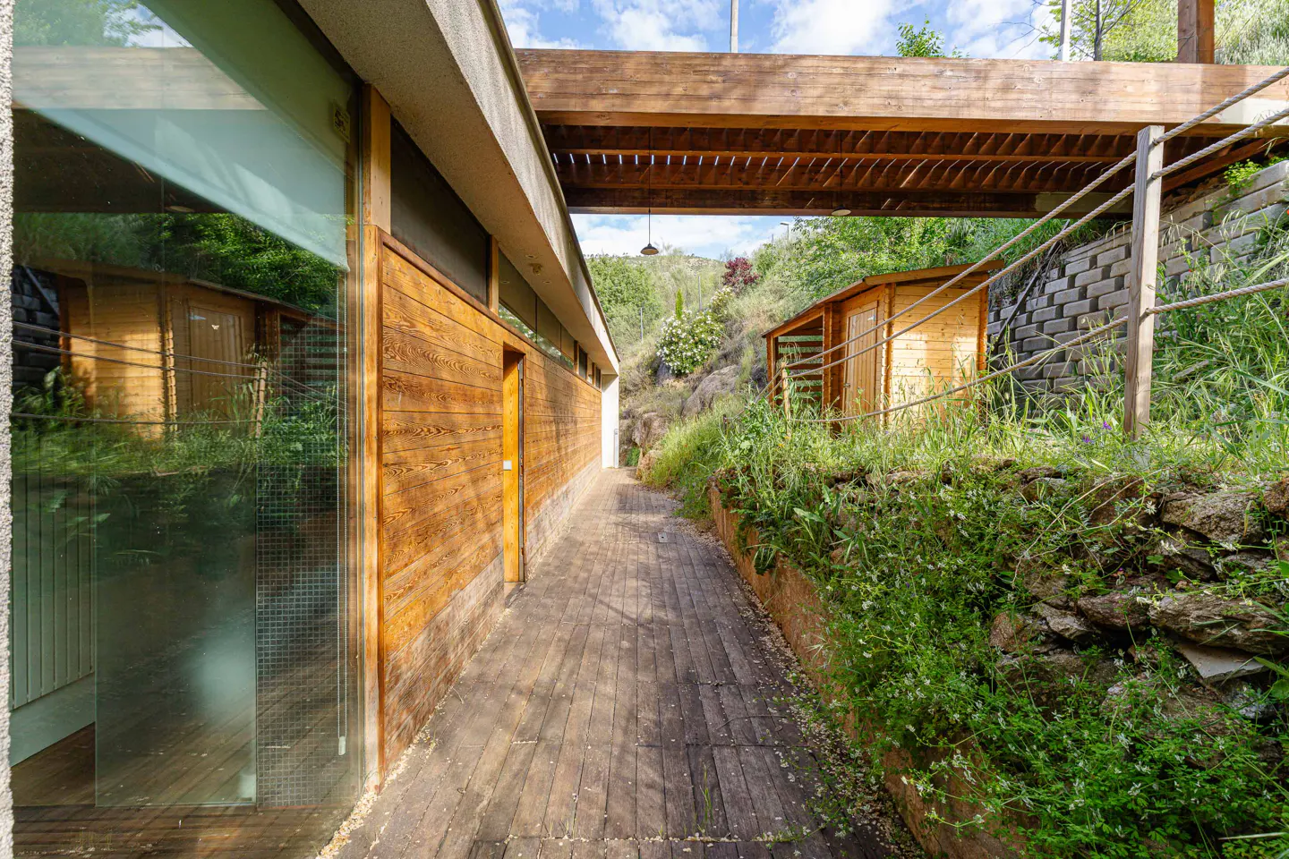 Wooden walkway with a building on the left and a shed on the right, surrounded by greenery.