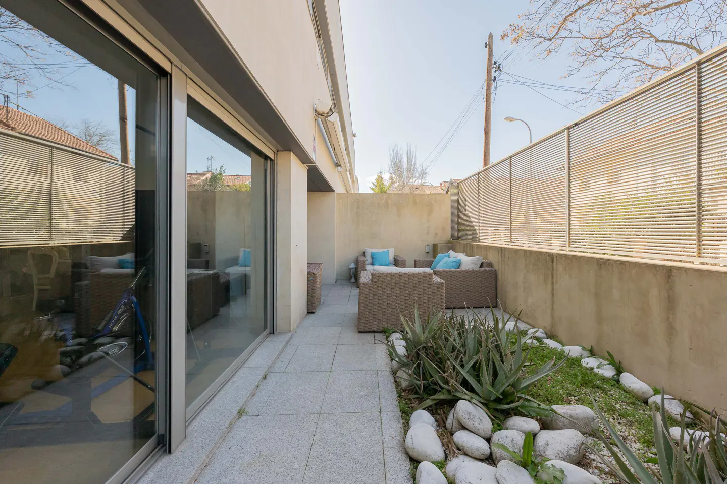 Outdoor patio with wicker furniture, blue pillows, and a rock garden. Sliding glass doors lead inside. A metal fence provides privacy.