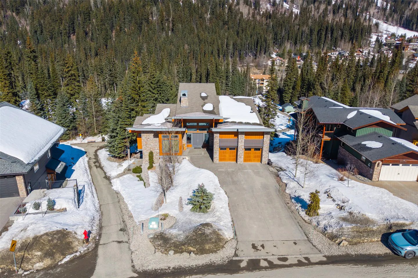 Aerial view of a modern home with a stone and wood facade, a driveway, and snow on the roof, surrounded by evergreen trees.