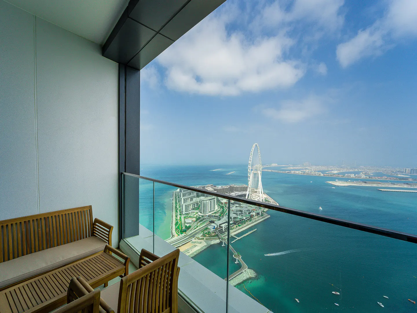 High-rise balcony with wooden furniture overlooking the ocean and Ain Dubai Ferris wheel on a sunny day.