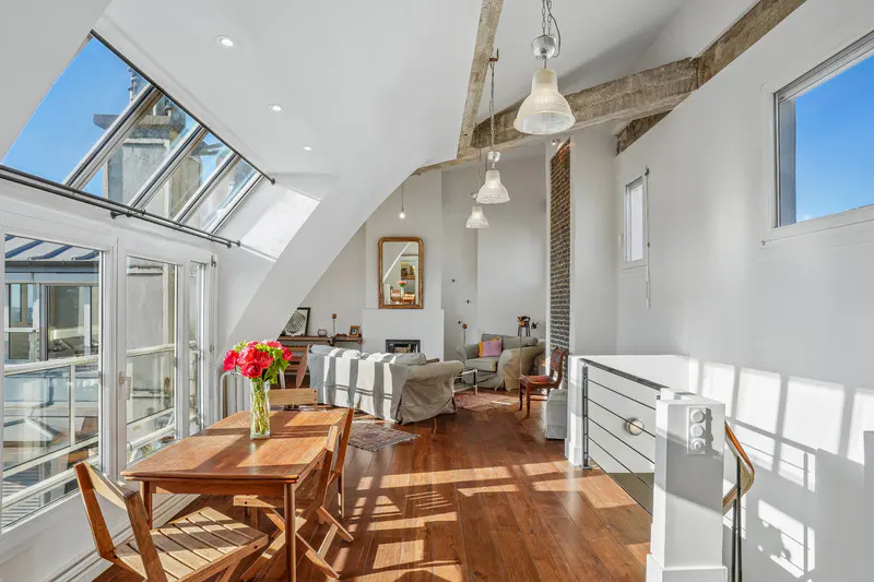 Bright, airy loft apartment with wood floors, white walls, and skylight. Dining table with flowers in foreground, living area with fireplace in background.