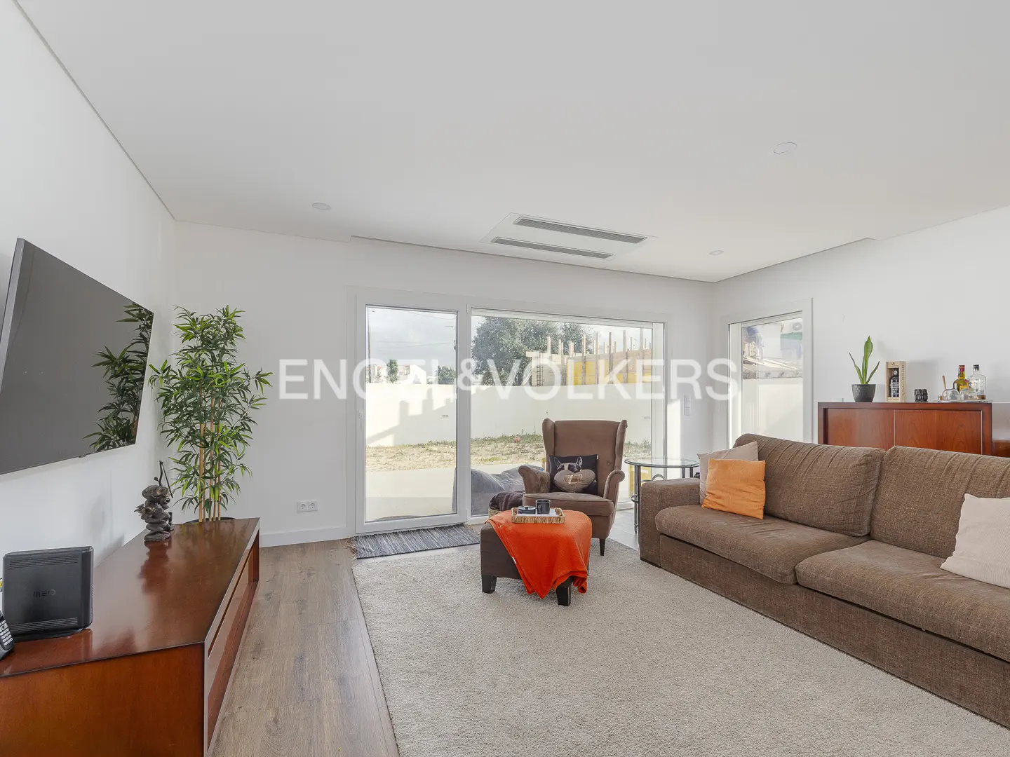 Bright living room with brown sofa, armchair, and TV. Large windows offer a view of the outdoors. Neutral carpet and white walls create a modern feel.
