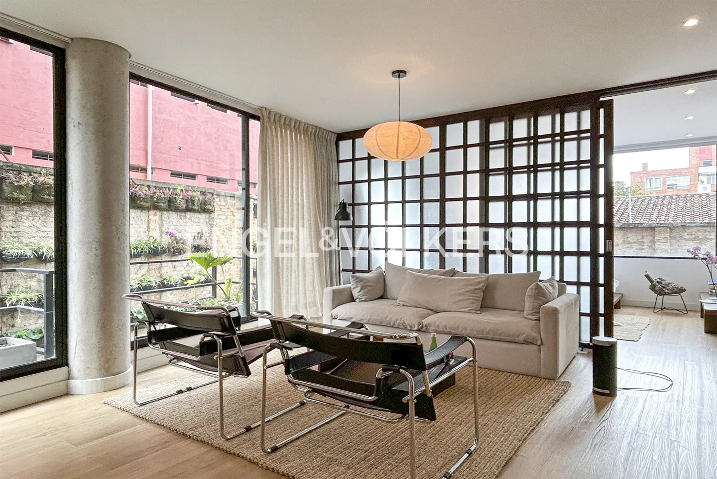 A bright living room with a beige sofa, two black chairs, and a large window with plants outside.