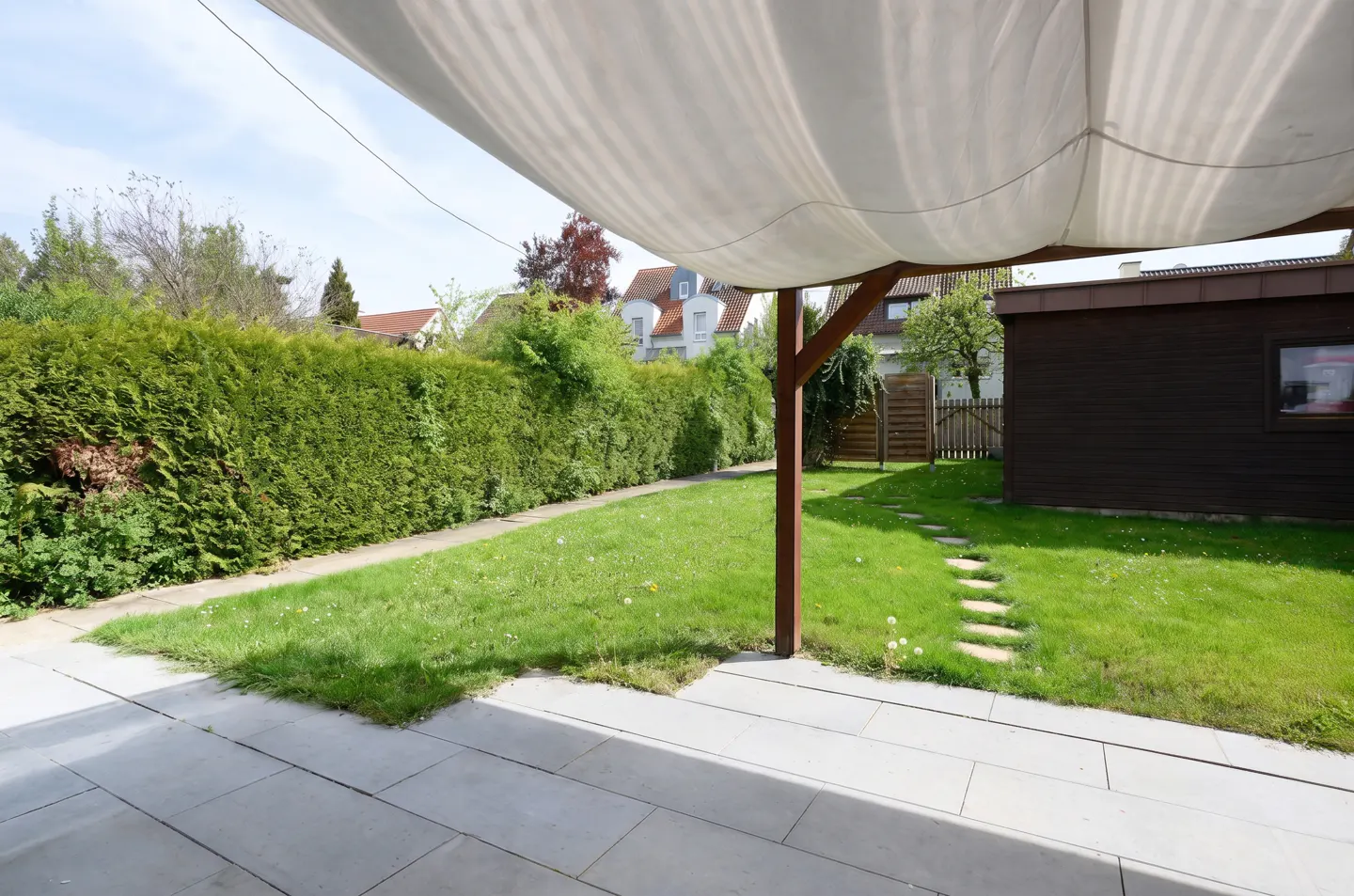 Backyard patio with gray stone tiles, green lawn, tall hedge, and a brown shed. A white awning provides shade.