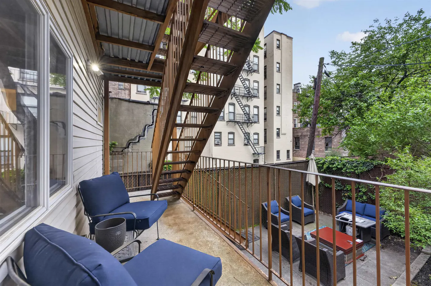 Outdoor balcony with blue chairs and a wooden staircase, overlooking a patio with outdoor furniture and city buildings.